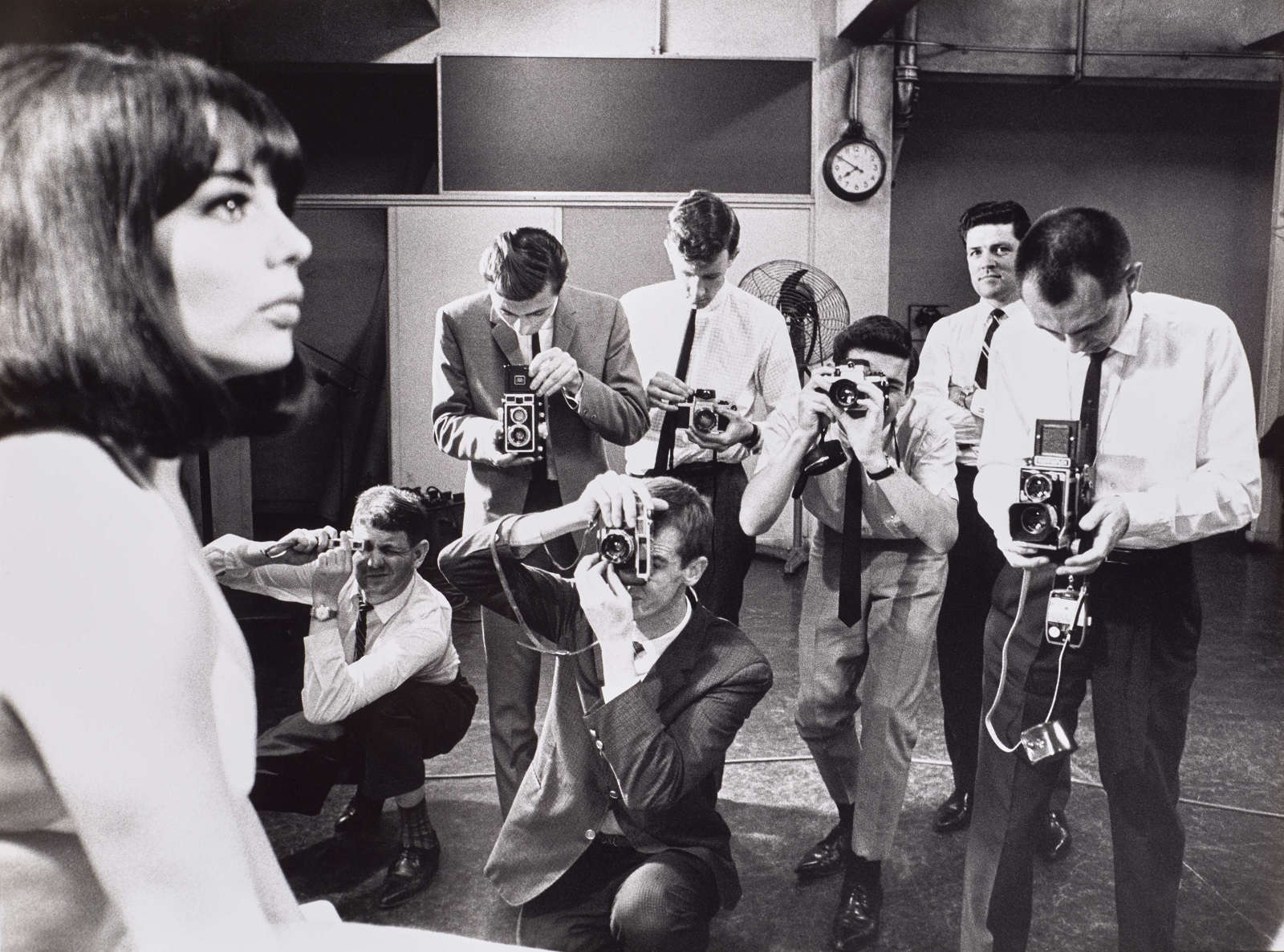 Seven male photography cadets dressed in collared shirts and ties photograph a female model in 1967