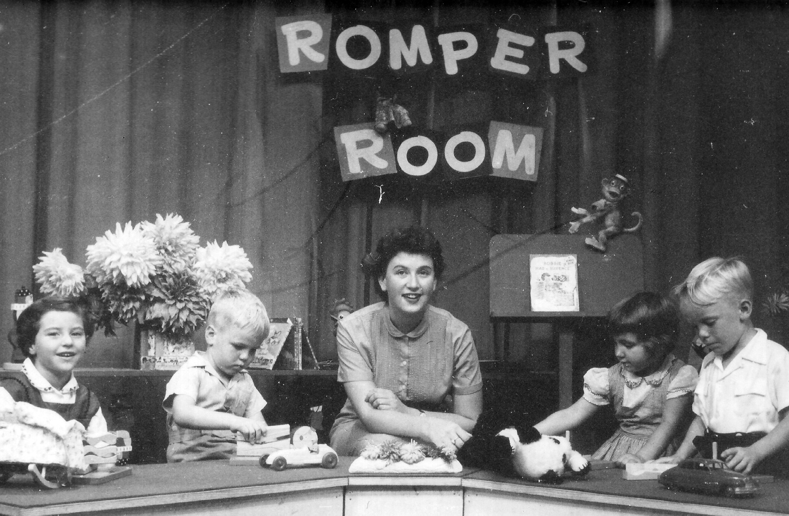 A woman sitting in the centre of a TV studio desk with children either side of her. A sign above her says 'Romper Room'. c1957.