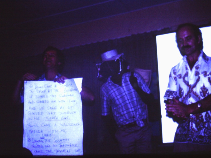 A photograph taken in a low-light setting that is blown out in some areas and too dark in others. Three figures appear to be singing and dancing. One man is holding up the words to 'Waltzing Matilda' on a piece of paper. Another wears a hat with corks.