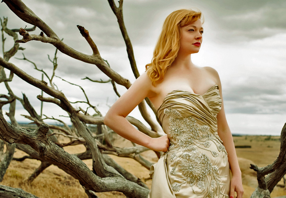 Still image of actress Sarah Snook wearing a strapless, champagne coloured, beaded couture gown in a scene from The Dressmaker, with an open dusty plain and gnarled trees in the background.
