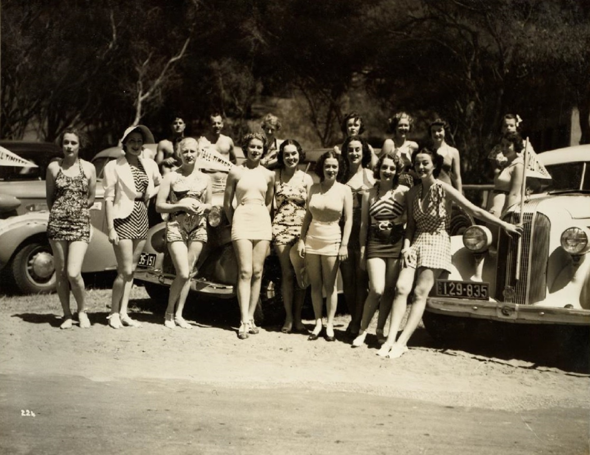 Men and woman in swimsuits lined up against cars for a photo.