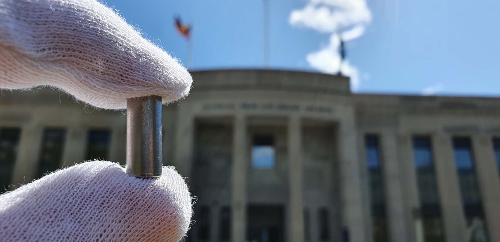 A gloved hand holding a small vial containing synthetic DNA, against the backdrop of the NFSA building in Canberra