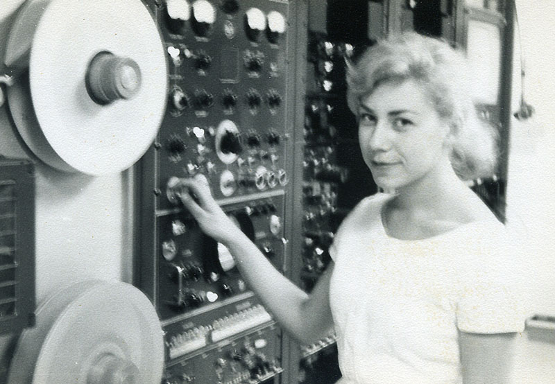 A woman is standing at a deck of videotape machines. She is operating one of the dials as she looks at the camera.
