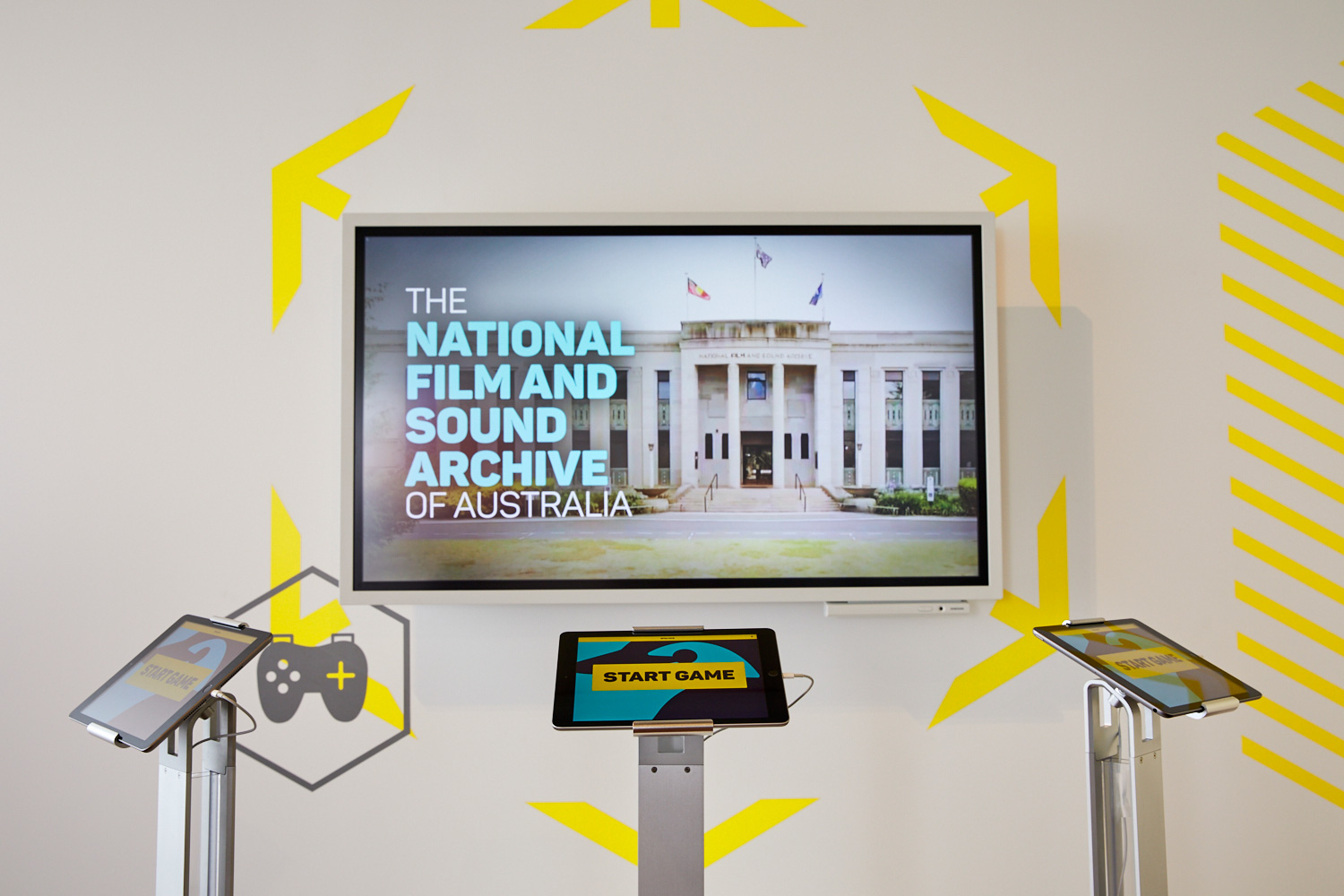 A large wall-mounted screen shows text saying 'The National Film and Sound Archive of Australia'. There are three ipads on stands in front of it displaying the graphics for the NFSA trivia game.