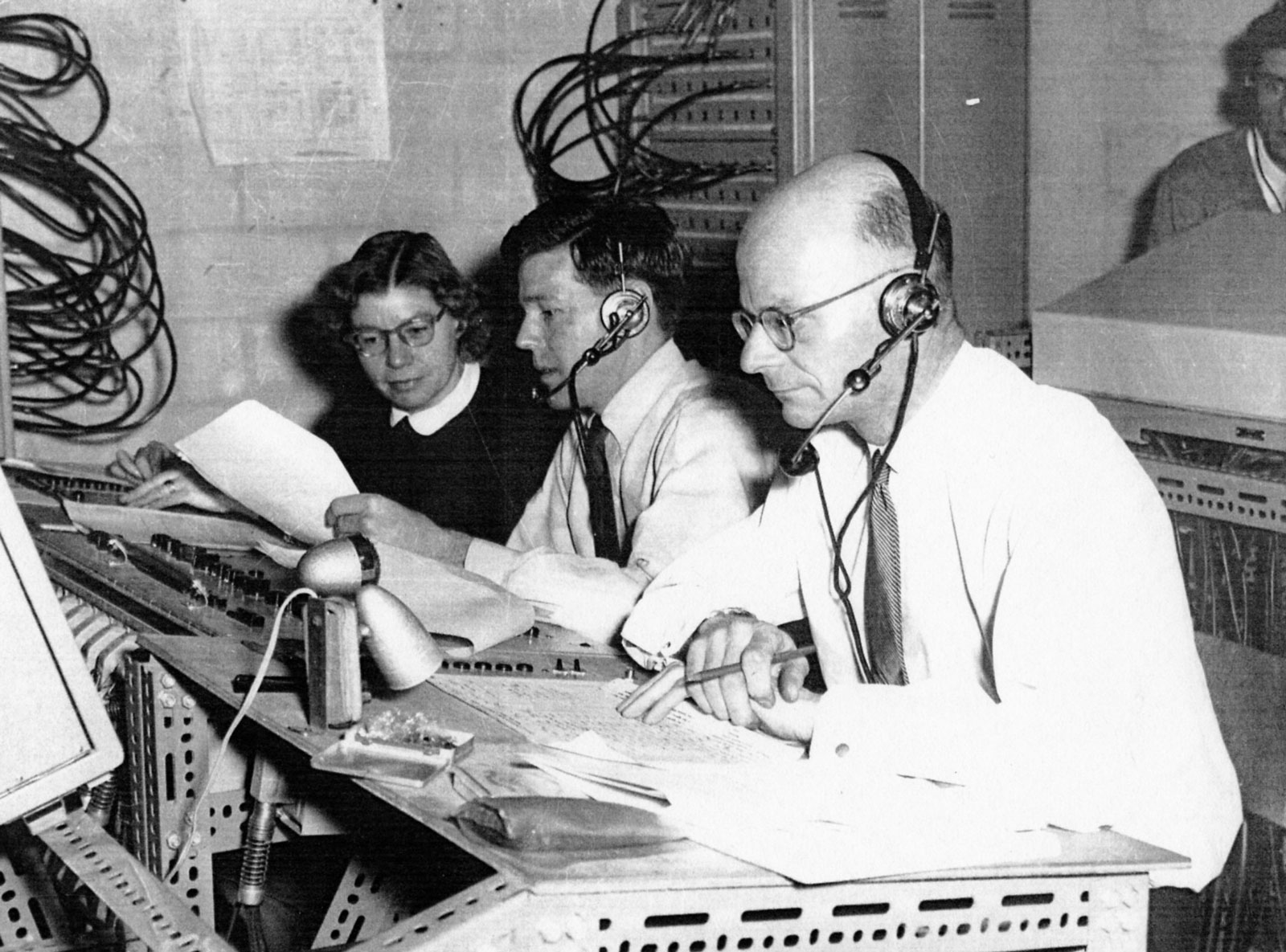 Two men and a woman sit behind a control desk in a black and white photo. There are lots of wires around them. The men wear headsets with headphones.