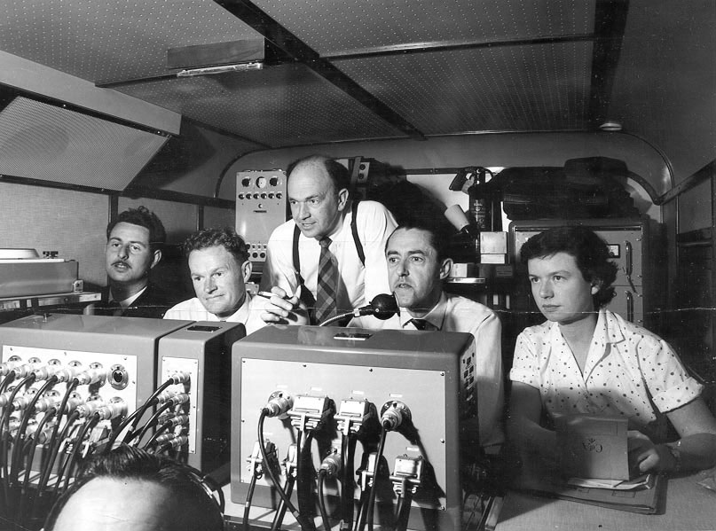 Four men and a woman sit inside an outside broadcast van. There are dials, cords and switches all around them.