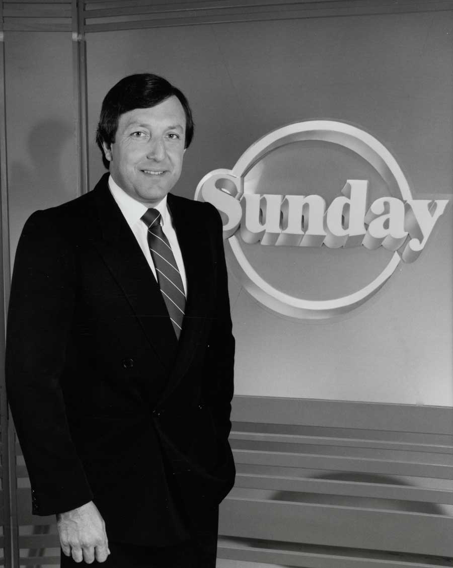 Journalist Jim Waley is wearing a suit and tie in this black and white photo. He is standing in a studio with the Sunday logo behind him.
