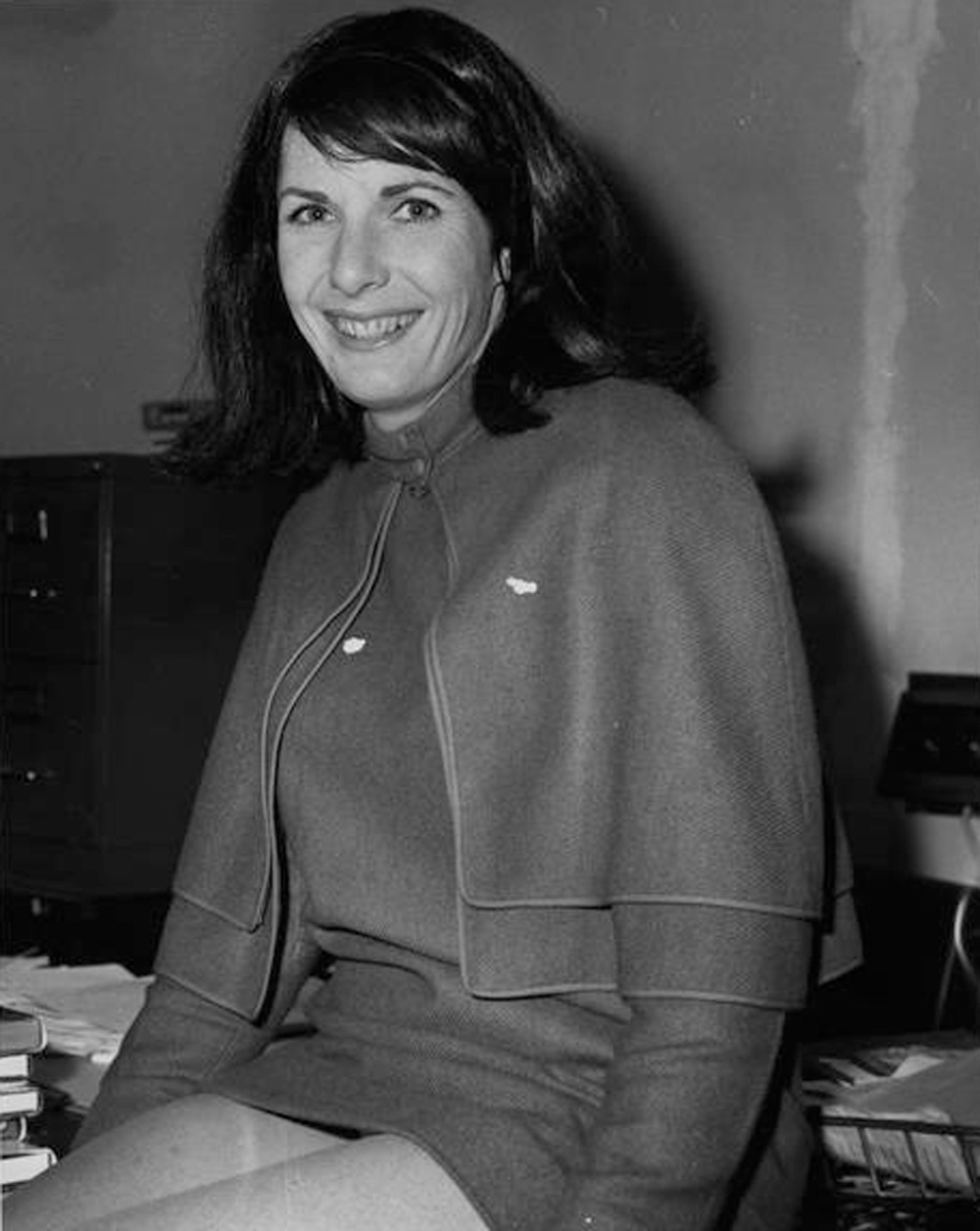 A woman is sitting on a desk and smiling at the camera.