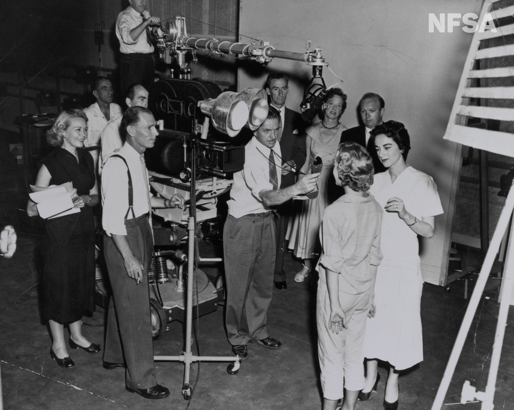 A group of people are involved in filming something with a large camera, lighting and sound. A woman is in front of the camera and another woman does her make-up. The crew is made up of men and women.