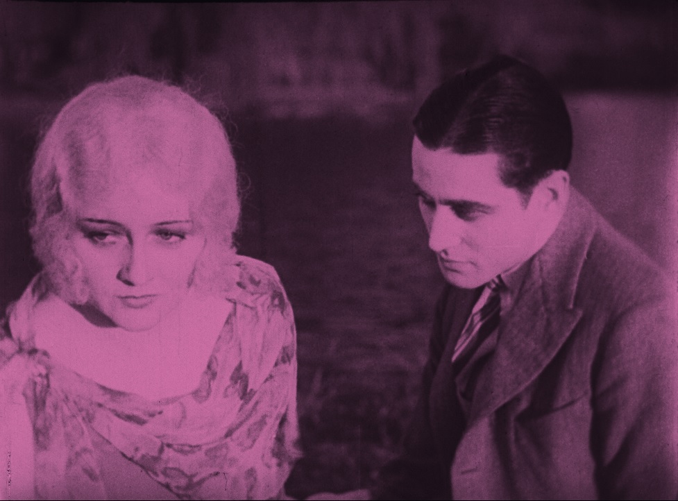 Pink tinted frame capture from the film, The Cheaters, shows aman and woman sitting by a lake 