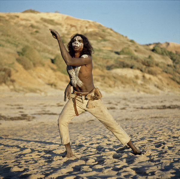 David Gulpilil as Fingerbone Bill in a production still from Storm Boy. He is striking a dance pose standing on a beach and painted with white paint on his face and torso