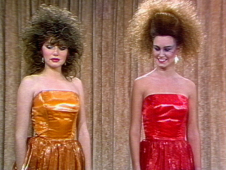 Two models are dressed in formal dresses - one orange and one bright pink. They have very big hair styles and lots of 80s-style make-up.