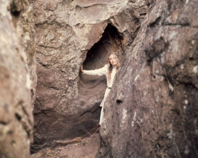 Anne Louise Lambert as Miranda, wearing a white dress and looking up through a gap in the rock in a scene from Picnic at Hanging Rock. 