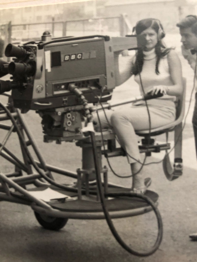 A woman is operating a BBC news camera on a rigging. She is wearing headphones. A man behind her peering into the camera viewfinder.