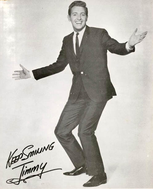 Black and white photograph of TV personality and singer Jimmy Hannan in a suit and tie. He has his arms outstretched and a big smile on his face. It is signed 'Keeping smiling, Jimmy'.