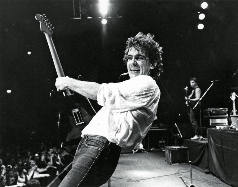 Guitarist Ian Moss from the band Cold Chisel on stage during a concert, smiling and leaning backward as he holds his guitar upright. 