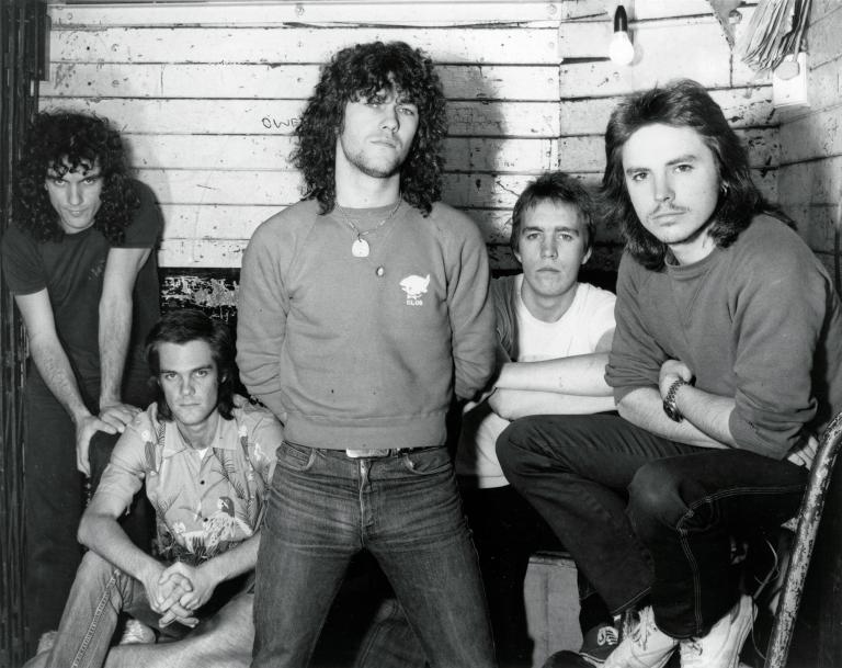 Members of the band Cold Chisel sitting and standing together in a wooden building, from left to right: Ian Moss, Don Walker, Jimmy Barnes, Steve Prestwich and Phil Small. 
