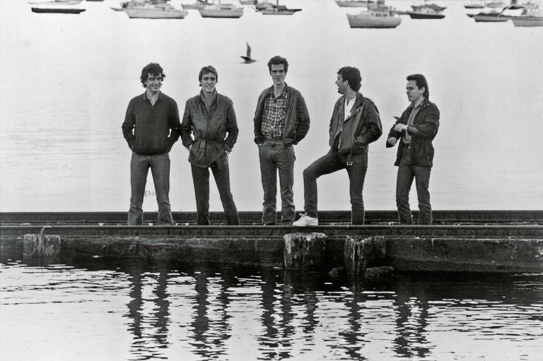Publicity photo of band Cold Chisel standing on a pier with boats in the distant background. From left to right: Ian Moss, Don Walker, Jimmy Barnes, Steve Prestwich, Phil Small.