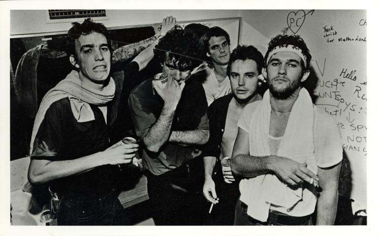The five band members of Cold Chisel in a graffitied room drinking beer and smoking cigarettes. From left to right: Steve Prestwich, Ian Moss, Don Walker, Phil Small, Jimmy Barnes.