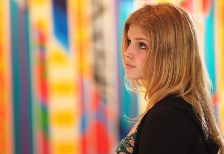 A young woman looking to her left is standing in front of colourful artwork in the background