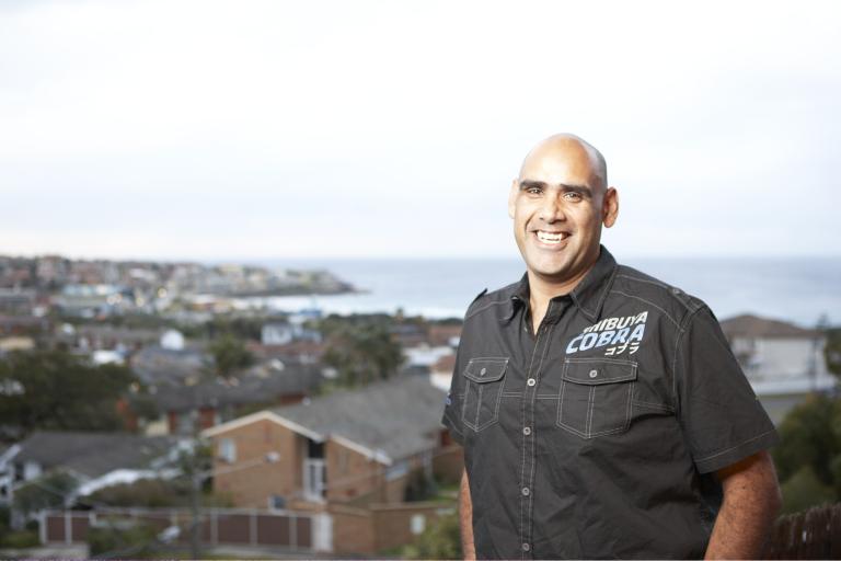 A man in a black shirt smiles in front of a suburban view with the sea in the distance