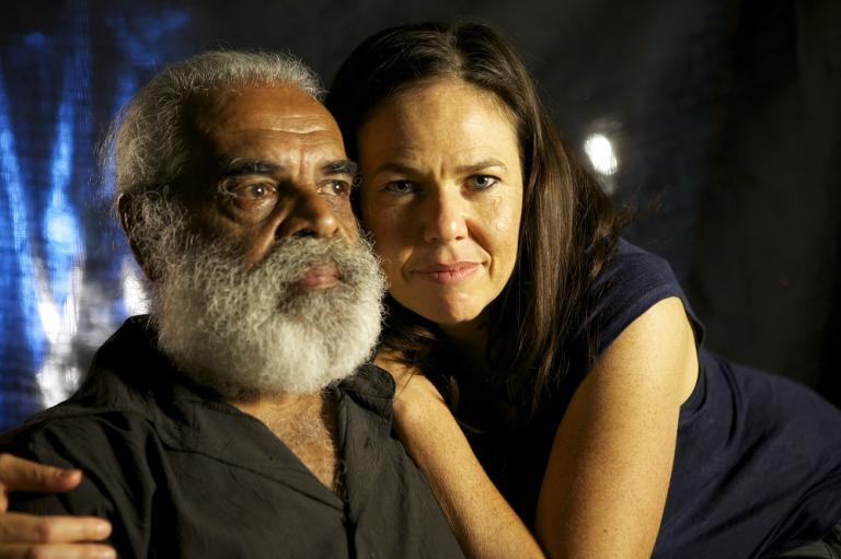 A woman leans on a bearded man and stares at the camera.