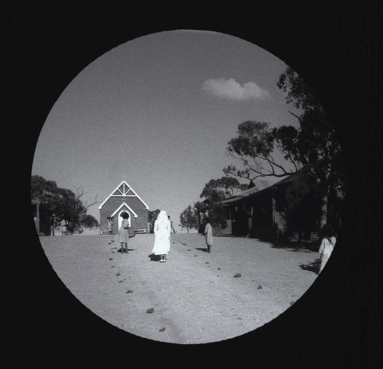 Black-and-white glass slide of a nun with several Indigenous children and a chapel in the background.