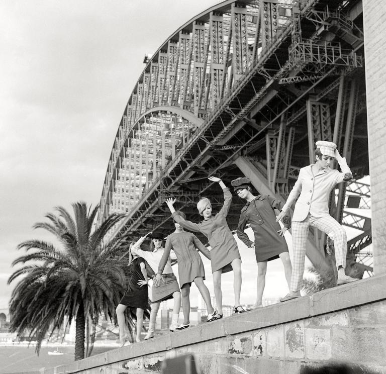 Black and white photo of six female models dressed in fashion from 1964 posing underneath the Sydney Harbour Bridge