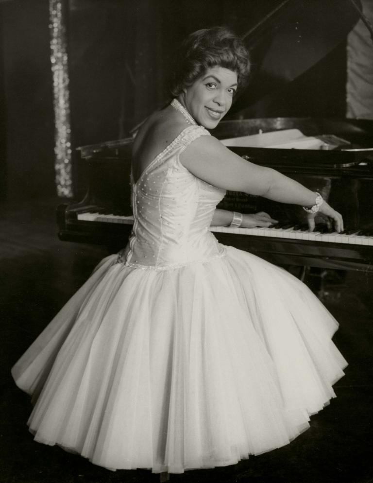 Publicity shot of Winifred Atwell seated at the piano and turning to face the camera