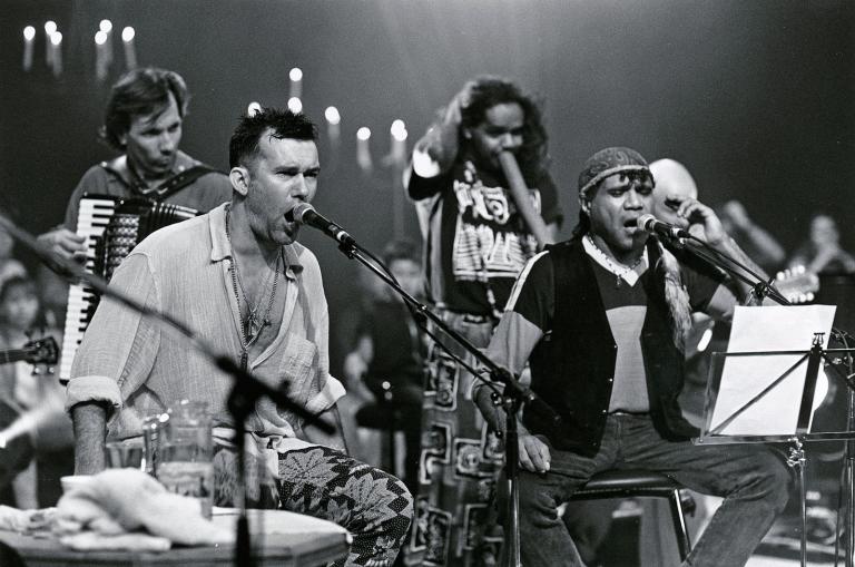 Singers Jimmy Barnes and Archie Roach on stage with their band during a live concert. Both are seated and singing into microphones.