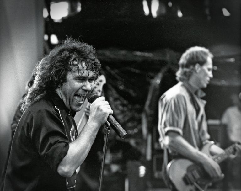 Jimmy Barnes performing on stage during one of the Australian Made concerts, singing into a microphone, with a guitarist in the background.