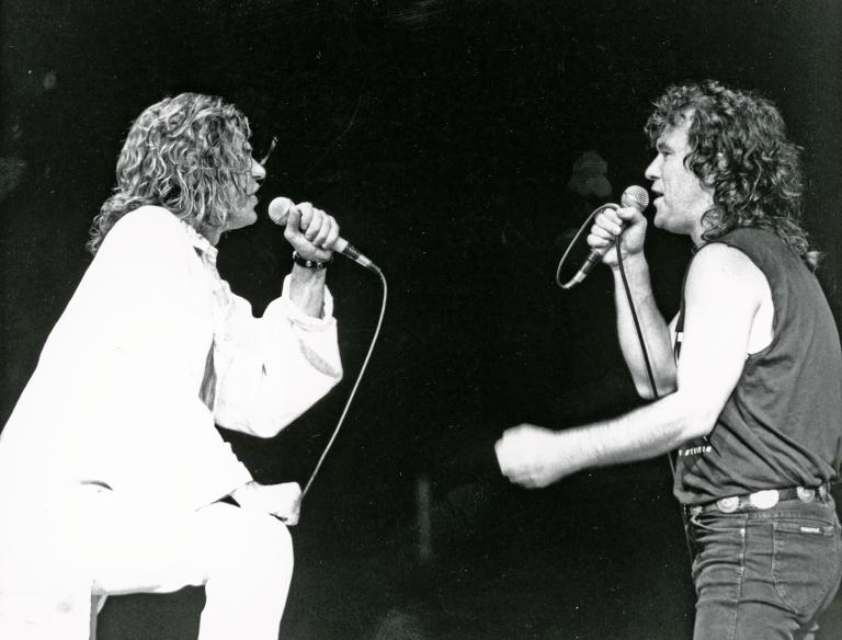 Close-Up of Michael Hutchence and Jimmy Barnes during a live concert, they are both holding microphones and facing each other on stage.