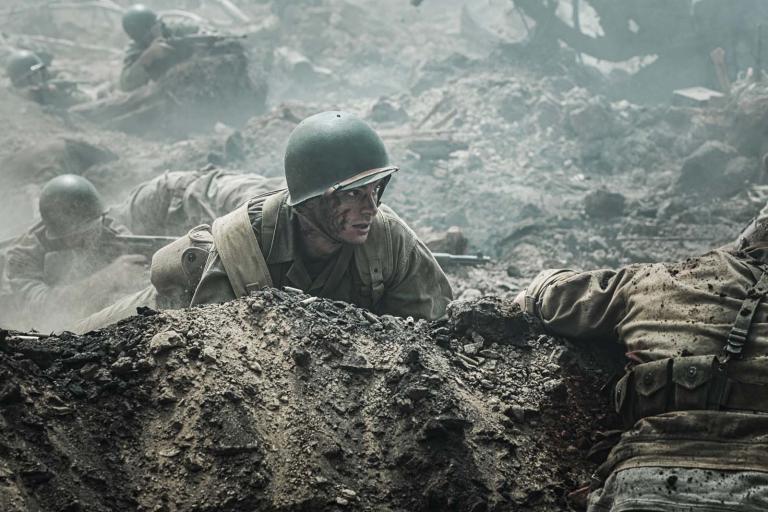 Andrew Garfield crouches behind a dirt mound with soldiers taking aim behind him and a soldier's body in the foreground, during the Battle of Okinawa in the Second World War