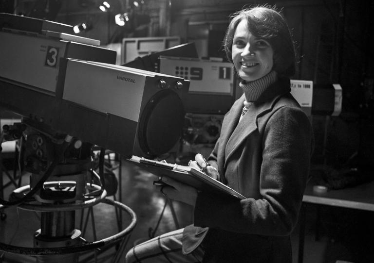 A woman holding a clipboard standing in a TV studio behind some TV cameras and studio lights.