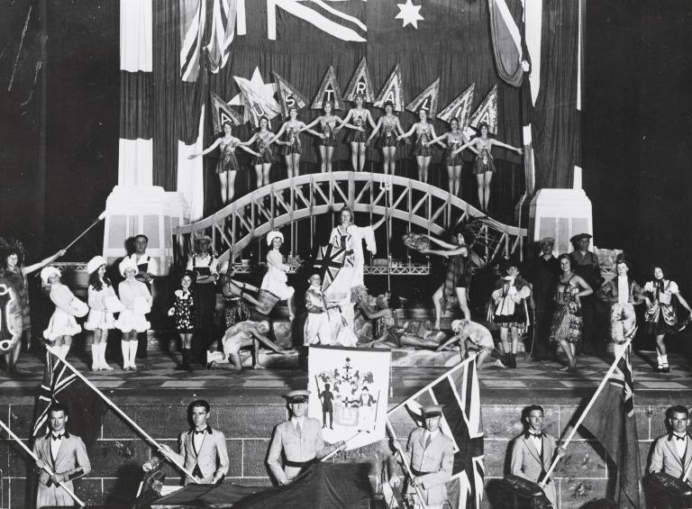 Large group pf people, in assorted costumes, on a stage in front of a small replica bridge. There are dancing ladies on top of the bridge and a line of men, in uniforms and suits, in the foreground holding flags.