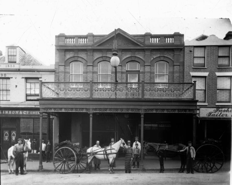 Men and children standing outside the Higgins Butcher in Hobart. Three horses and two carts stand outside. The building has intricate ironwork decoration.