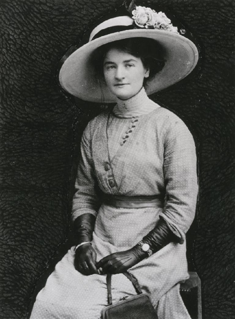 Seated photo of Ella Caspers with handbag, gloves and large hat with flowers.
