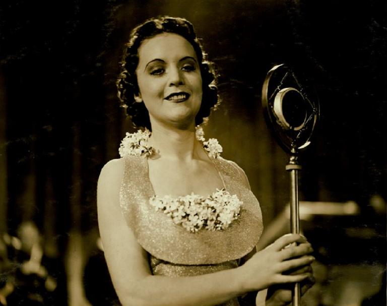 A woman wearing a dress woth flowers around the neckline stands at a microphone