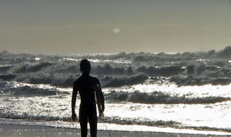 A silhouette of a man at the beach walking away from camera into the surf carrying a surfboard under one arm. 