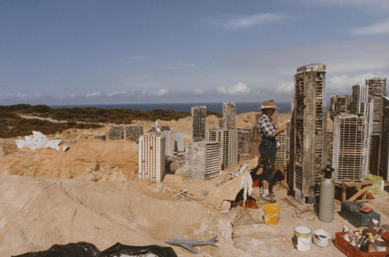 A crew member constructing a model set of post-apocalyptic Sydney in some sand dunes. The buildings are about the size of the person. The Opera House is visible. 