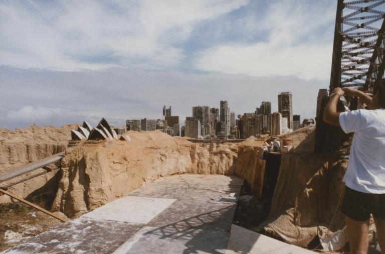 A crew member next to a model of the Sydney Harbour Bridge on the set of post-apocalyptic Sydney in some sand dunes. The Opera House and city skyline is also visible.