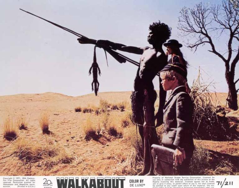 David Gulpilil stands pointing at something in the distance. The stick he uses to point has a dead goanna hanging off it. Next to him are two white school children.