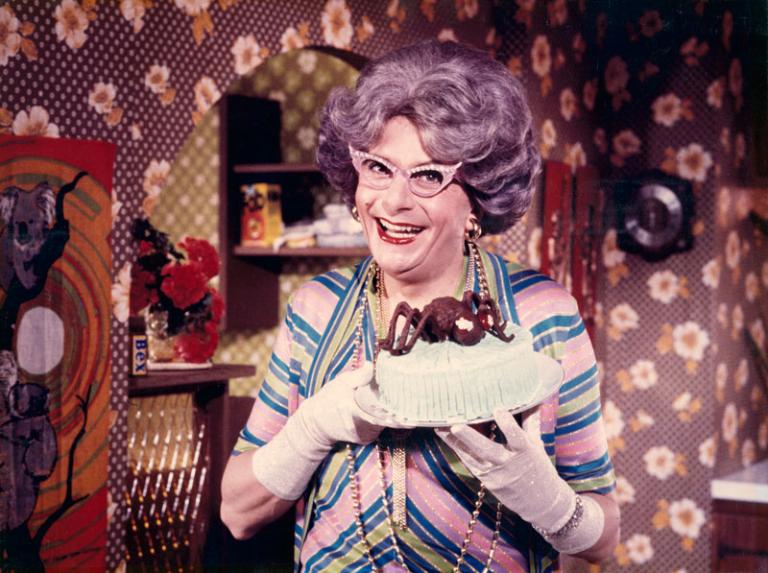 Dame Edna Everage is standing in a brightly coloured room. She has a mauve bouffant hairdo and her trademark winged glasses. She is holding an iced cake with a chocolate spider on top with gloved hands.