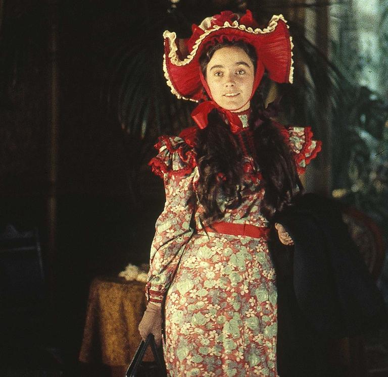 A young woman dressed in a period floral-patterned dress with a red belt and an ostentatious red, broad-brimmed hat