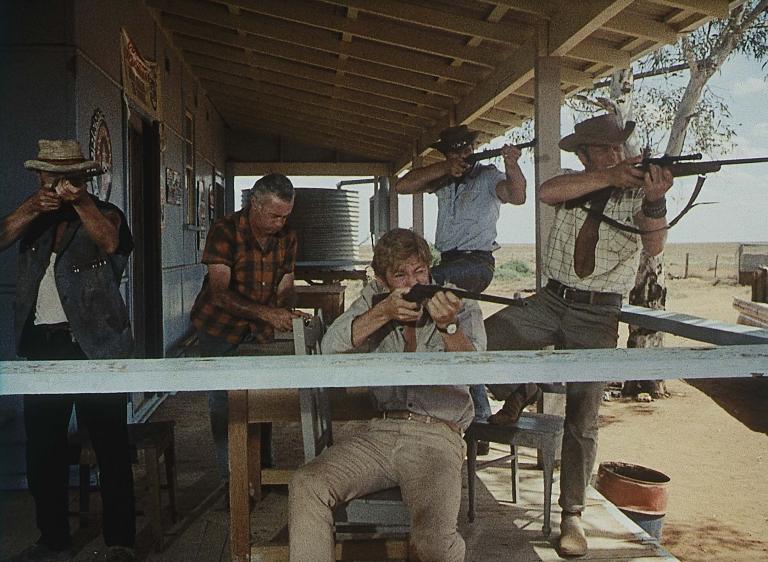 Five men on the balcony of an outback house, four of whom are pointing rifles in the same direction