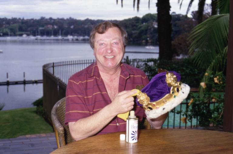 Graham Kennedy sitting at a table smiling to camera holding his crown with a bottle of Brasso in front of him