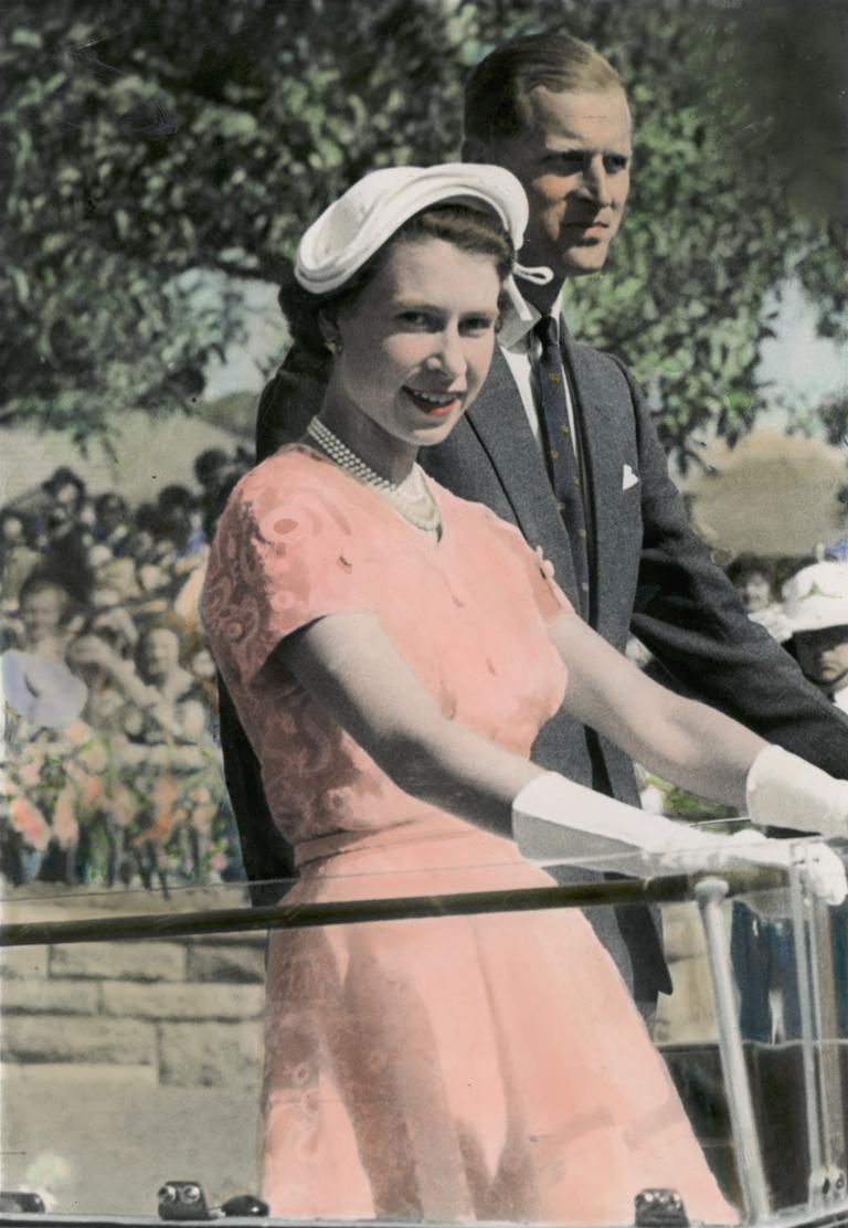 Queen Elizabeth II and Prince Philip in 1954. The Queen is wearing a pink frock with white hat and gloves, holding onto a rail and Philip is standing behind her wearing a suit.