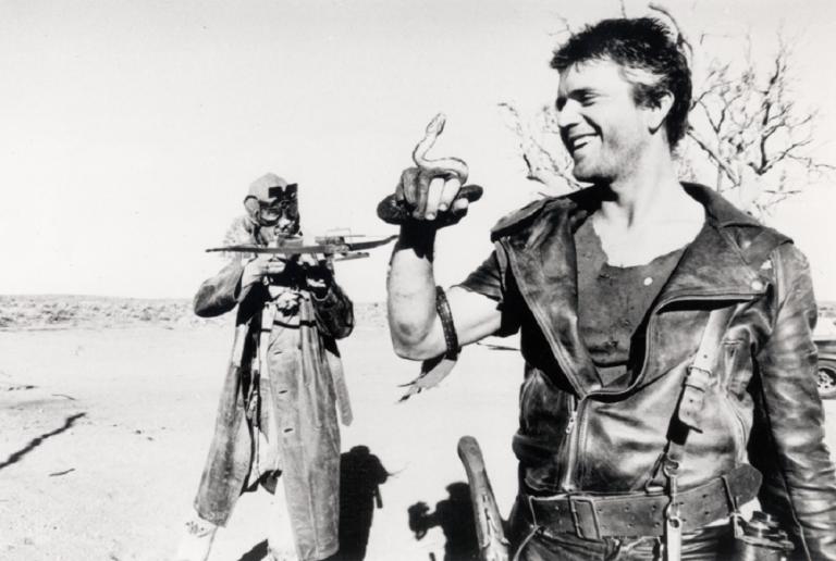 Max (Mel Gibson) holds a snakes while the Gyro Captain (Bruce Spence) points a crossbow at him