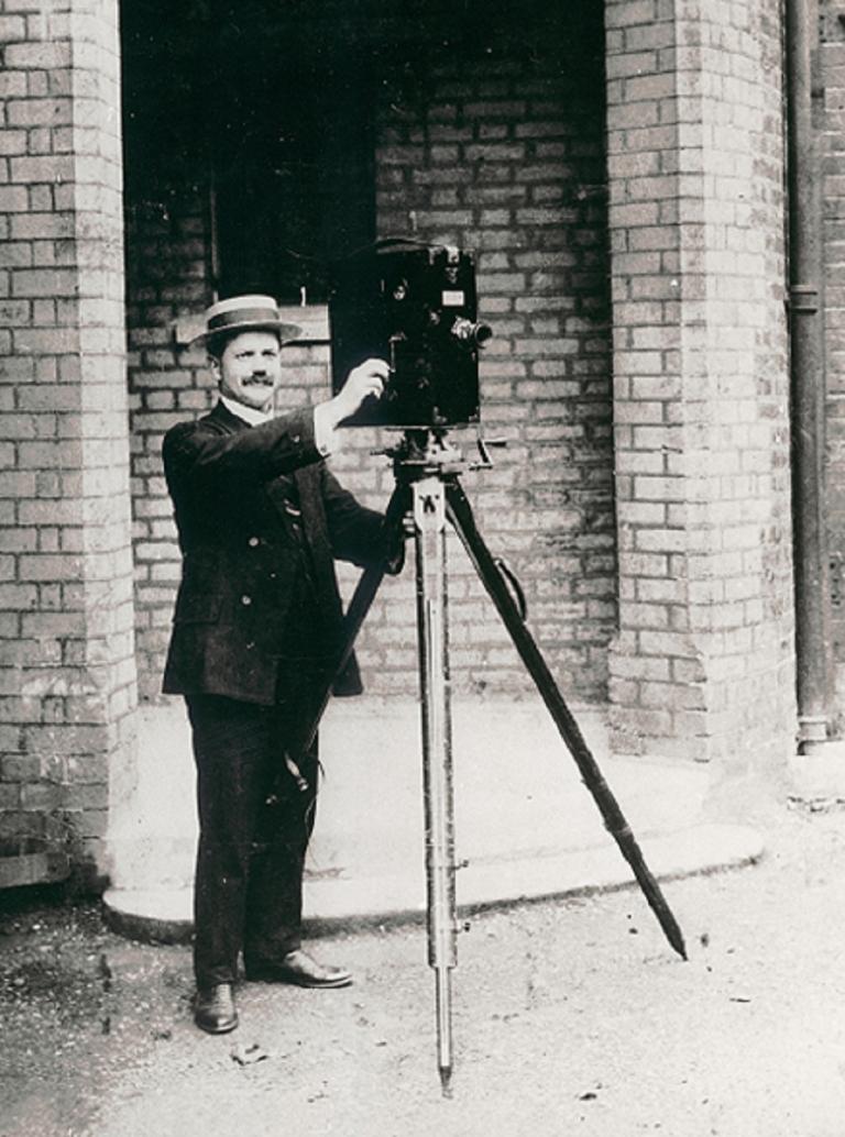 Sidney Cook standing outside a building with his movie camera mounted on a large tripod.