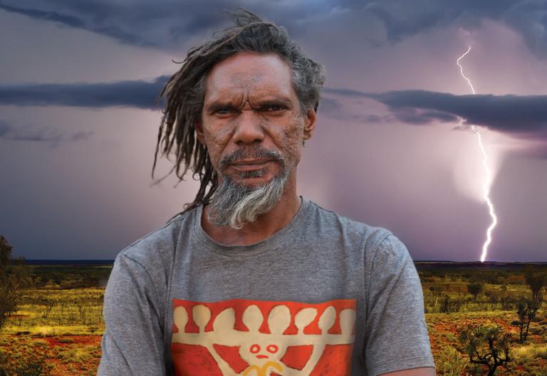 A bearded, middle-aged Aboriginal man in the outback stands staring at camera. In the distant background you can see lighting striking the ground.